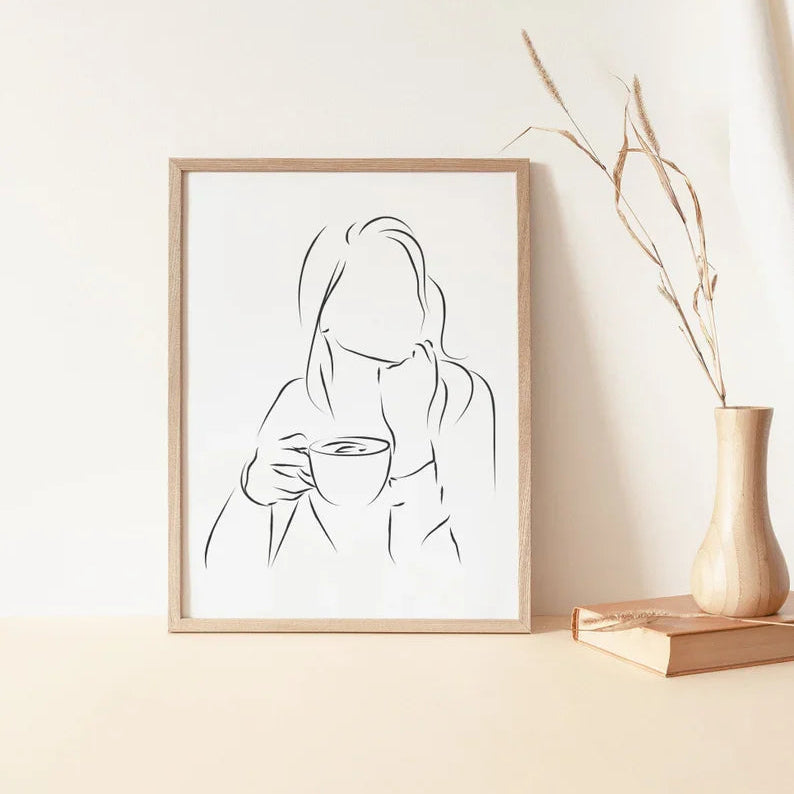 A framed line art drawing of a woman holding a mug, displayed on a wooden surface with a boho frame.