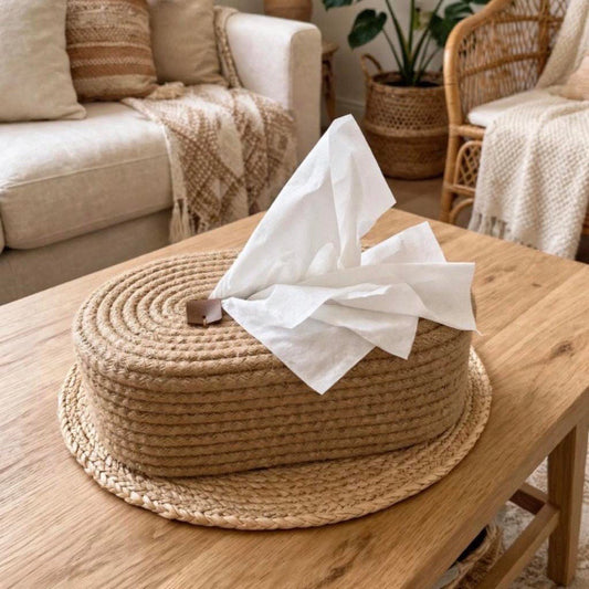 A round woven straw hat-shaped tissue box cover or decorative holder sits on a wooden coffee table in a cozy living room. White tissue paper is tucked into the top opening.