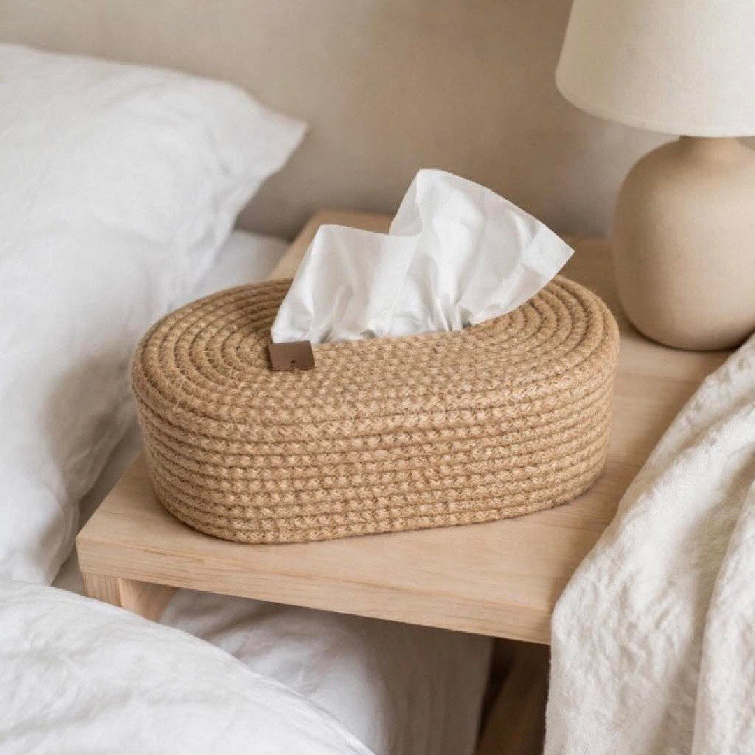 A woven oval tissue box holder in natural jute sits on a light wood bedside table next to a bed with white bedding and a beige table lamp.